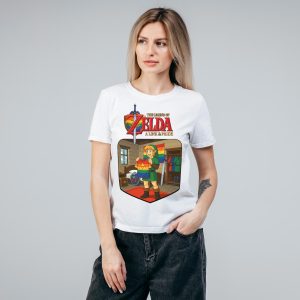 Une femme aux cheveux blonds portant un tee-shirt blanc orné d'un graphique coloré de The Legend of Zelda : A Link to Pride, célébrant la communauté LGBTQIA+, se tient sur un fond gris uni, une main dans la poche.