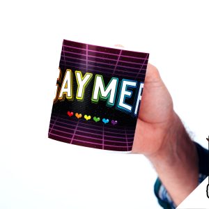 Une personne vêtue d'une chemise à carreaux tient une tasse sur laquelle on peut lire GAYMER en lettres colorées avec des cœurs arc-en-ciel en dessous. Dans le coin, un logo BREAK THE CLOSET représentant une tête de licorne sur fond d'arc-en-ciel est parfait pour tout joueur queer fier de l'être.