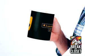 Une personne tient une tasse noire avec des symboles partiels jaunes et blancs ; le dessin n'est pas clair. Dans le coin, un logo "Break the Closet" avec une licorne et un arc-en-ciel représente fièrement la communauté LGBTQIA+.