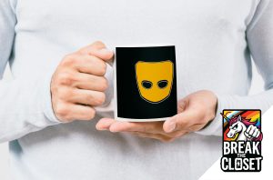 Une personne vêtue d'une chemise blanche tient un mug noir portant le logo d'un masque jaune. Dans le coin, un graphique "Break the Closet" (Brisez le placard) avec des couleurs arc-en-ciel, un texte en gras et une licorne, célèbre la fierté queer et la visibilité des LGBTQIA+.