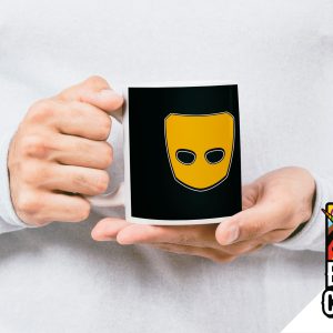 Une personne vêtue d'une chemise blanche tient un mug noir portant le logo d'un masque jaune. Dans le coin, un graphique "Break the Closet" (Brisez le placard) avec des couleurs arc-en-ciel, un texte en gras et une licorne, célèbre la fierté queer et la visibilité des LGBTQIA+.