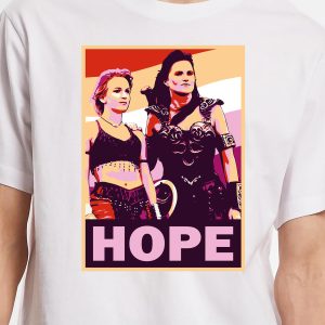 Un t-shirt présente un graphique stylisé et coloré de deux femmes guerrières queer se tenant côte à côte au-dessus du mot "HOPE" en lettres grasses. L'arrière-plan présente des nuances vibrantes d'orange, de rose et de crème.