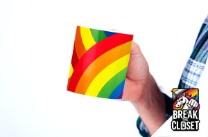 Une personne portant une chemise à carreaux tient une tasse avec un motif arc-en-ciel. Dans le coin inférieur droit, un logo sur le thème des LGBT représente une licorne et le texte BREAK THE CLOSET sur fond noir.