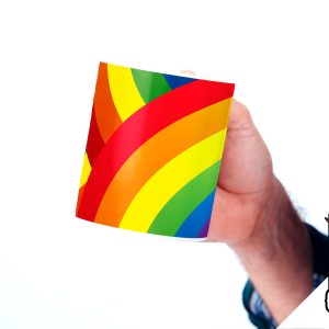 Une personne portant une chemise à carreaux tient une tasse avec un motif arc-en-ciel. Dans le coin inférieur droit, un logo sur le thème des LGBT représente une licorne et le texte BREAK THE CLOSET sur fond noir.
