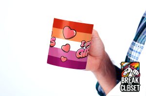 Une personne tient une tasse décorée de cœurs et de rayures horizontales aux couleurs du drapeau de la fierté lesbienne, avec un texte disant I prefer girls (Je préfère les filles). Un logo "Break the Closet" représentant une licorne se trouve en bas à droite, célébrant la fierté LGBTQIA+.
