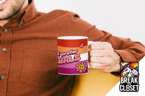 Une personne vêtue d'une chemise de couleur rouille tient une tasse avec un drapeau de la fierté lesbienne, des cœurs, une fleur et les mots "I prefer girls" (je préfère les filles). Le logo BREAK THE CLOSET avec un arc-en-ciel et une licorne célèbre la fierté LGBTQIA+ dans le coin inférieur droit.