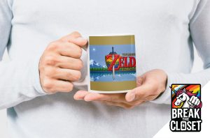 Une personne vêtue d'une chemise claire tient une tasse arborant le logo de Legend of Zelda : A Link to the Past ; dans le coin, il y a un logo Break the Closet avec une licorne et un poing levé - célébrant la fierté queer.