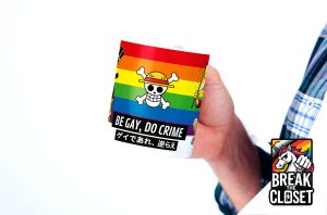 Une personne tient une tasse avec un drapeau arc-en-ciel, un crâne de pirate dans un chapeau de paille et le texte BE GAY, DO CRIME en caractères japonais. Le logo de la licorne "BREAK THE CLOSET" apparaît dans le coin de l'image.