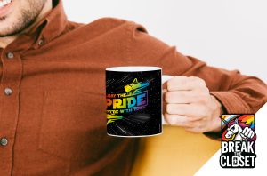 Une personne vêtue d'une chemise de couleur rouille sourit en tenant une tasse noire avec une licorne arc-en-ciel et les mots "May the PRIDE be with you" (que la fierté soit avec vous). Dans le coin, un logo "Break the Closet" célèbre la fierté LGBTQIA+.