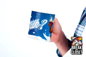 Une main tient une tasse bleue avec le mot Milk ? et l'illustration d'une personne aspergée de lait. Dans le coin, un logo coloré avec une licorne et le texte BREAK THE CLOSET célèbrent la fierté LGBTQIA+.