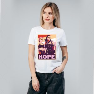 Une femme aux longs cheveux blonds porte un T-shirt blanc sur lequel figure un graphique de style pop art représentant deux femmes, avec le mot HOPE en lettres grasses en dessous - un clin d'œil élégant à la fierté LGBTQIA+ - sur un fond gris clair uni.