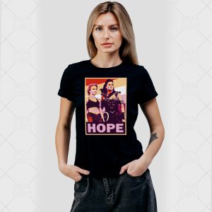 Une femme aux longs cheveux blonds, vêtue d'un t-shirt noir orné d'un graphique pop art coloré représentant deux femmes homosexuelles et le mot HOPE en dessous d'elles, se tient debout sur un fond clair uni, les mains dans les poches.