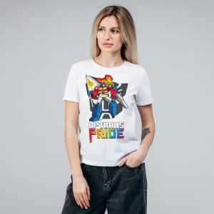 Une personne aux longs cheveux blonds portant un t-shirt blanc représentant un robot coloré tenant un drapeau de la fierté et le texte "Optimus Pride" dans les couleurs de l'arc-en-ciel, se tient sur un fond gris uni, célébrant fièrement l'identité queer et LGBT.