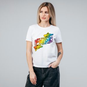 Une femme aux longs cheveux blonds porte un t-shirt blanc avec des vaisseaux spatiaux aux couleurs de l'arc-en-ciel et le texte May the Pride be with you, célébrant la fierté LGBTQIA+. Elle se tient debout, une main dans la poche, sur un fond gris.