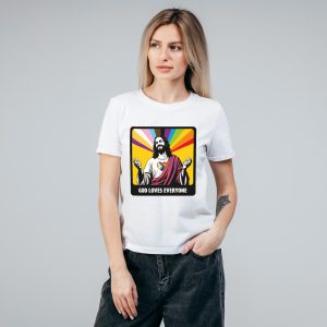 Une femme aux longs cheveux blonds porte un t-shirt blanc représentant Jésus avec des rayons arc-en-ciel et le texte GOD LOVES EVERYONE, exprimant son soutien à la communauté LGBTQIA+. Elle se tient debout sur un fond gris uni, une main dans la poche.