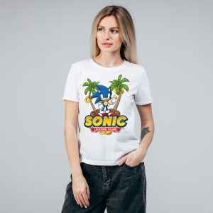 Une femme portant un T-shirt blanc avec un graphique coloré de Sonic le hérisson, des palmiers et des anneaux d'or célèbre la fierté homosexuelle. Le texte sur le tee-shirt indique SONIC COCKERING ISLAND. Elle se tient debout sur un fond gris uni.
