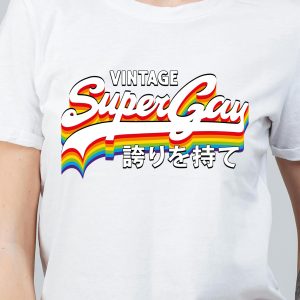 Une personne portant un t-shirt blanc avec les mots Vintage Super Gay en lettres arc-en-ciel et un texte en japonais en dessous, célébrant fièrement l'identité LGBTQIA+ sur un fond uni.