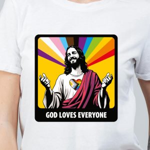 Un T-shirt présente un personnage illustré ressemblant à Jésus avec un cœur arc-en-ciel sur la poitrine, des rayons colorés en arrière-plan et le texte GOD LOVES EVERYONE en dessous de l'image - une célébration audacieuse de la fierté LGBTQIA+.