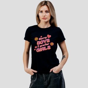 Une femme aux longs cheveux blonds porte un t-shirt noir sur lequel est écrit "Sorry boys, I prefer girls" en lettres roses et rouges avec des motifs de cœurs et de fleurs, exprimant fièrement son identité queer sur un fond uni.