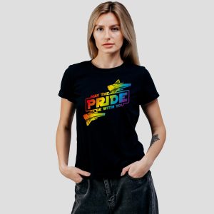 Une femme se tient debout sur un fond uni, portant un T-shirt noir sur lequel on peut lire "May the PRIDE be with you" dans les couleurs de l'arc-en-ciel avec deux vaisseaux spatiaux, célébrant fièrement la fierté LGBT. Elle a les cheveux clairs et une expression faciale neutre.