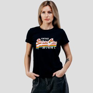 Une femme aux longs cheveux blonds porte un t-shirt noir sur lequel on peut lire Vintage Super Gay avec des rayures arc-en-ciel et des caractères japonais, célébrant la fierté homosexuelle. Elle se tient debout sur un fond gris clair uni, les mains dans les poches.