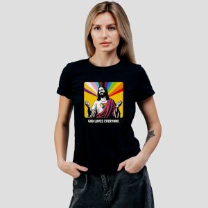 Une femme portant un t-shirt noir avec un graphique coloré de Jésus, un fond arc-en-ciel et le texte God Loves Everyone (Dieu aime tout le monde) - une déclaration audacieuse de fierté queer et LGBT - se tient sur un fond gris clair uni.