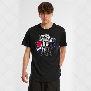 Un jeune homme portant un T-shirt noir avec un graphique indiquant Leather Pride Team au-dessus de deux personnages illustrés en tenue de cuir, debout devant un arrière-plan de fierté LGBTQIA+ rayé bleu, blanc, rouge et noir.