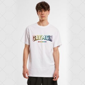 Une personne se tient debout, portant un t-shirt blanc sur lequel est inscrit GAYMER en lettres arc-en-ciel, avec une rangée de petits cœurs arc-en-ciel en dessous - un fier clin d'œil aux communautés LGBT et queer. L'arrière-plan est uni et de couleur claire.