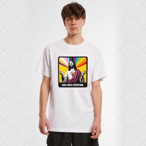 Un jeune homme portant un T-shirt blanc avec un graphique coloré de Jésus, des rayons arc-en-ciel en arrière-plan et le texte "God Loves Everyone" (Dieu aime tout le monde) - un clin d'œil fier à la communauté LGBTQIA+.