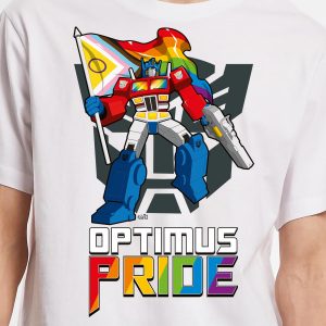 Optimus Prime tient un drapeau Progress Pride devant un symbole Autobot. En dessous, on peut lire OPTIMUS PRIDE avec PRIDE dans les couleurs de l'arc-en-ciel, célébrant la fierté LGBTQIA+ sur un T-shirt blanc.