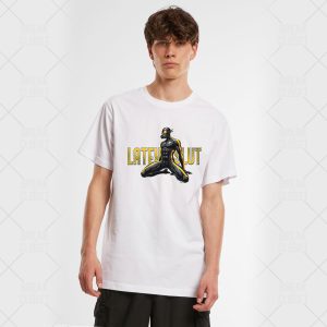 Un jeune homme homosexuel aux cheveux bruns bouclés porte le T-shirt unisexe Latex Slut, qui présente un super-héros masqué agenouillé et un texte jaune en gras "LATE PLUT". Il se tient sur un fond clair et uni.