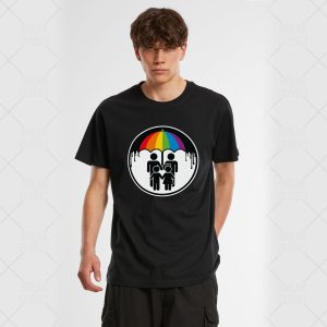 Un jeune homme aux cheveux bruns ondulés porte un t-shirt noir représentant deux personnages sous un parapluie coloré, célébrant la fierté LGBTQIA+, avec de la peinture noire dégoulinant de la bordure circulaire autour du motif.