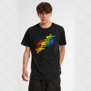 Une personne portant un t-shirt noir avec un texte aux couleurs de l'arc-en-ciel indiquant May the Pride Be With You, avec des stries arc-en-ciel et un graphique de vaisseau spatial, se tient sur un fond uni - une célébration audacieuse de la fierté queer et LGBTQIA+.