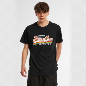 Un jeune homme queer aux cheveux bruns ondulés porte un T-shirt noir sur lequel est inscrit Vintage Super Gay en lettres rétro colorées, avec un petit arc-en-ciel et un texte en japonais en dessous, sur un fond blanc uni.