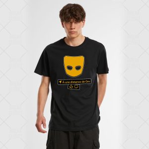Un jeune homme aux cheveux ébouriffés porte un t-shirt noir orné d'un grand masque jaune et d'un texte en français indiquant À une distance de 0m avec de petites icônes en dessous, ce qui constitue un clin d'œil subtil à l'identité homosexuelle. Il se tient debout sur un fond clair.